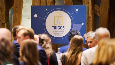 Im Vordergrund befinden sich mehrere Personen, teilweise unscharf abgebildet. Im Zentrum hängt ein blaues Schild mit dem TRIGOS Logo und dem Text „TRIGOS – Die Auszeichnung für verantwortungsvolles Wirtschaften“