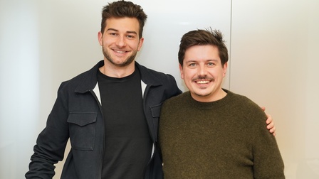 Zwei Personen stehen nebeneinander vor einem hellen Hintergrund und blicken lächelnd in die Kamera. Eine Person hat den Arm um die andere gelegt. Beide tragen legere Oberbekleidung und stehen dicht beieinander.