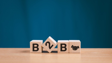 Auf einem Tisch aus hellem Holz sind vier Würfel platziert, wobei der zweite schief liegt, die anderen sind aufgestellt. Auf den Würfel steht B2B und am vierten Würfel ist das Symbol von sich schüttelnden Händen. Der Hintergrund ist blau