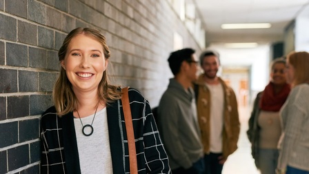 Halbporträt einer Person mit hellen, offenen Haaren, die sich an einer dunklen Ziegelmauer anlehnt. Um die linke Schulter hat sie eine Umhängetasche. Im Hintergrund verschwommen stehen vier Personen in einem Kreis.
