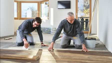 Zwei Personen in grauen Arbeitsmonturen knien am Boden und verlegen Holzbodenbretter, im Hintergrund Fenster, Balkontüre und Fernseher