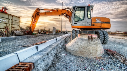 Ein Bagger steht auf einer Baustelle einer Straße. Links neben dem Bagger ist eine Erhöhung für einen Gehsteig. Neben dem Bagger liegen Metallstäbe
