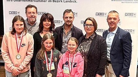 Eine Gruppe von acht Personen steht vor einem Hintergrund mit dem Logo der Wirtschaftskammer Tirol. Vorne stehen drei Kinder mit Medaillen um den Hals; zwei tragen Nike-Pullover. Hinter ihnen stehen fünf Erwachsene – drei Männer und zwei Frauen – in festlicher, aber alltäglicher Kleidung. Alle blicken lächelnd in die Kamera. Die Atmosphäre wirkt wie eine Preisverleihung oder Ehrung im Bezirksverband Landeck.