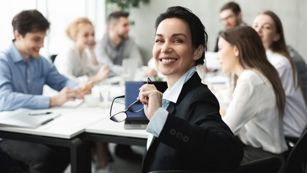 Person mit dunklen geschlossenen Haaren und Businesskleidung hält eine Brille in der Hand und blickt freudig in die Kamera, während im Hintergrund weitere Personen am selben Besprechungstisch sitzen