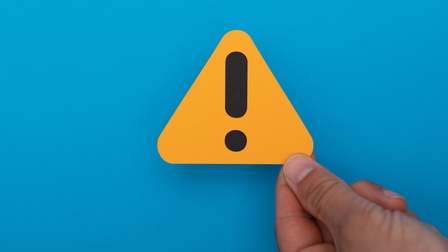 Man sieht das Symbol eines gelben Dreiecks mit einem schwarzen Rufzeichen auf blauem Hintergrund. Das Icon wird von einer Hand gehalten und symbolisiert Gefahr. 