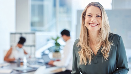 Person mit langen blonden Haaren und dunkelgrüner Bluse lächelt freudig in die Kamera, im Hintergrund sitzen Personen an einem Schreibtisch in Unschärfe und arbeiten