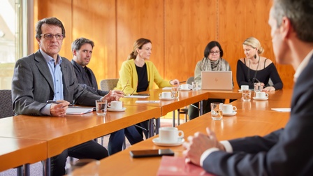 Personen sitzen an Tischen in U-Form und nehmen an einer Besprechung teil, eine Person in Anzug mit Brille blickt gespannt zu einer Person gegenüber