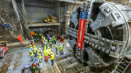 Foto einer unterirdischen Baustelle aus erhöhter Perspektive. Rechts nimmt der große, kreisförmige Schneidkopf einer Tunnelbohrmaschine einen wesentlichen Teil des Bildes ein. An der Maschine hängt ein rot-blaues Banner mit der Aufschrift „WIENER LINIEN“. Links neben der Maschine steht eine Gruppe von Personen mit Schutzhelmen und Warnwesten