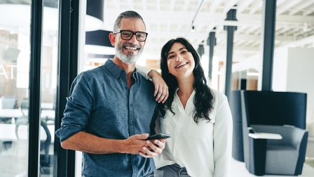 Eine lächelnde Person mit langen schwarzen Haaren legt Hand auf Schulter einer anderen bärtigen Personen