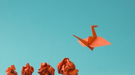 Ein orangefarbener Origami-Vogel aus Papier schwebt vor einem einfarbig blauen Hintergrund. Am unteren Bildrand liegen mehrere zerknüllte Papierkugeln aus demselben Material