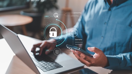 Person sitzt vor einem Laptop bei einem Schreibtisch und hält ein Handy in der linken Hand. Über Handy und Laptop schweben verschiedene Symbole wie ein Schloss in einem Kreis oder eine Weltkugel mit einem Pfeil