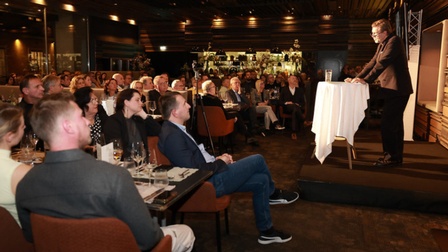 Eine größere Gruppe von Menschen hört einem Vortragenden zu, der auf einer kleinen Bühne an einem Stehtisch spricht.