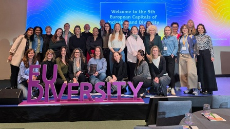 Mehrere Personen stehen und knien in zwei Reihen auf einer Bühne. Im Vordergrund steht der Schriftzug „EU DIVERSITY“. Im Hintergrund ist eine große Projektionsfläche mit dem Text „Welcome to the 5th European Capitals of Inclusion and Diversity“