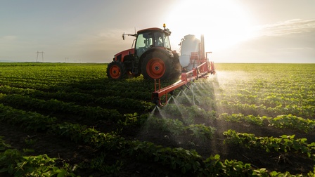 Ein roter Traktor mit breitem schwarzen Aufsatz mit Spritzdrüsen, der über Feld mit grünen Pflänzchen fährt und Flüssigkeit darauf sprüht.