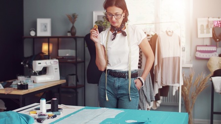 Person mit Brillen und Maßband um den Hals blickt auf mit Nadeln abgesteckte Schablonen am Tisch vor ihr liegend, ringsum Zwirne, im Hintergrund verschwommen Nähmaschine und Kleiderstange 