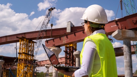 Eine Person ist von hinten fotografiert und hält einen Laptop in der Hand. Sie betrachtet die Metallkonstruktion einer Brücke, neben der sich auch ein Kran befindet. Am Himmel sind Wolken, es herrscht Sonnenschein.