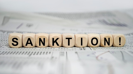 Neun Würfel liegen nebeneinander auf bedrucktem Papier. Auf jedem Würfel ist ein Buchstabe, wodurch sich das Wort Sanktion ergibt. Am letzten Würfel ist ein Rufzeichen