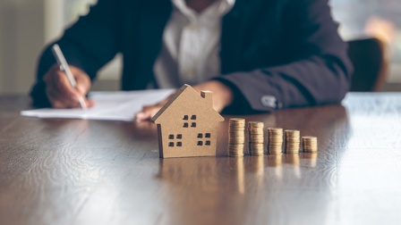 Auf einem Tisch steht ein kleines Modellhaus. Daneben stehen nebeneinander fünf, unterschiedlich hohe Münzstapel. An dem Tisch sitzt eine Person, vor der ein Zettel ist, auf dem die Person etwas notiert