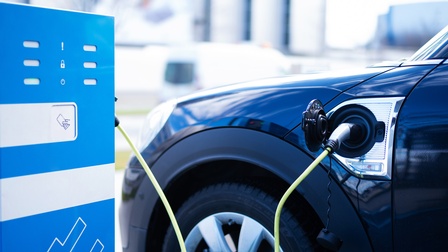 Seitliche Nahaufnahme des linken, vorderen Teils eines blauen Autos. Links neben dem Auto steht eine blaue Box, aus der ein grünes Kabel kommt. Beim Auto ist eine Klappe geöffnet. Das Kabel ist dadurch am Auto angesteckt.