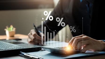 Eine Person sitzt an einem Tisch. Vor ihr steht ein aufgeklappter Laptop. In der rechten Hand hält sie einen Stift. Mit der linken Hand tippt sie auf einem Taschenrechner. Darüber ist grafisch eine Balkenstatistik gelegt sowie drei Prozentzeichen