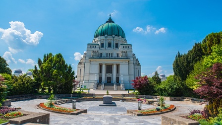 In der Mitte ist ein runder Platz mit einem steinern Sarg. Links und rechts davon sind grüne Büsche und Bäume. Neben dem Platz ist eine Aufbewahrungshalle mit einer grünen Kuppel