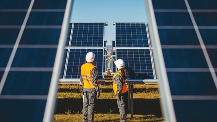 Rückenansicht zweier Personen mit Helmen und Sicherheitswesten, die auf einem Feld zwischen zwei Solarkollektoren stehen. Dahinter sind viele weitere Solarkollektoren