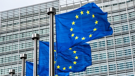 Drei EU-Flaggen im Wind wehend, blaue Stoffe mit gelben im Kreis verlaufenden Sternen an Fahnenmasten, im Hintergrund Hausfassade mit Sonnenblenden