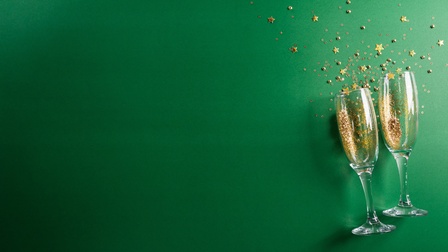 Auf grünem Untergrund liegen in der rechten Bildhälfte nebeneinander zwei  Gläser,. In den Gläsern sind goldene Partikel. Rund um die Gläser liegen kleine goldene Sterne in Form von Konfetti. 