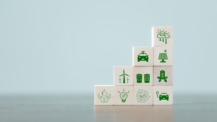 Mehrere Holzwürfel aufeinander gestapelt auf einer hellen Oberfläche mit grünen Symbolen zum Thema Klimaschutz: Eine Wolke mit der Aufschrift CO2, Autos, Solar-Panel, Windräder, Mistkübel, Glühbirne, Pfeile, Handflächen mit Wassertropfen.