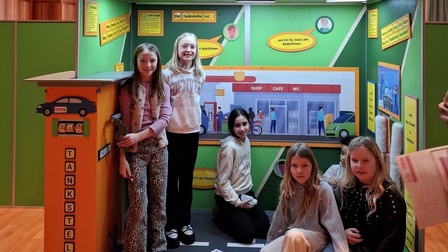 Mehrere Kinder befinden sich in einem thematisch gestalteten Innenraum mit dem Titel „Kinder-Erlebnis-Raum KiERa“. Der Raum ist wie eine Tankstelle gestaltet, mit einer Zapfsäule, Beschriftungen und einer illustrierten Wanddarstellung. Die Kinder stehen und sitzen innerhalb der Installation und blicken in die Kamera.