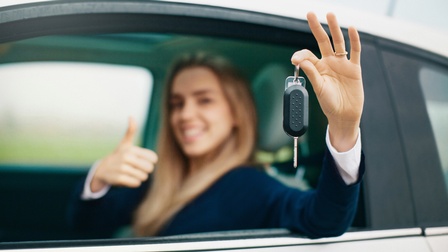 Nahaufnahme von einer einem Autoschlüssel, der von einer Hand gehalten wird, in der Unschärfe sitzt eine Person in einem Auto und zeigt mit dem Daumen nach oben