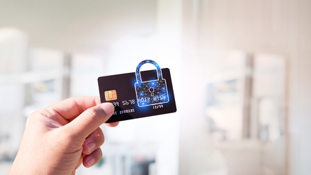 Nahaufnahme der Hand einer Person, in dieser hält sie eine Kreditkarte in der Kamera. Auf der Karte ist das Symbol eines Schlosses, der Verschluss ragt über die Karte. Im Hintergrund ist verschwommen eine Wohnung zu sehen