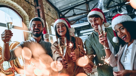 Frontale Aufnahme von vier Personen mit Weihnachtsmützen, die in einem Büroraum stehen und jeweils ein Sektglas und Sprühkerzen halten