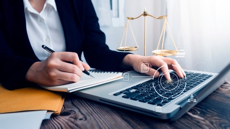 Topshot der Hände einer Person, die mit einem Kugelschreiber in ein Notizbuch schreibt. Vor ihr ist ein Laptop auf einem braunen Holztisch aufgeklappt. Man sieht grafische Symbole von  Personen, Notizen und einem Parlament. Neben ihr  steht eine Waage.