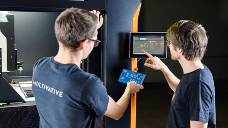 Zwei der Gründer vor der Maschine. Einer bedient eine Touch-Board, der andere hält eine blaue Platine