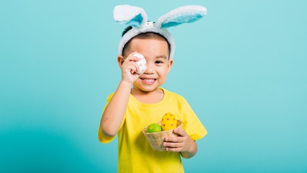 Halbporträt eines Kindes, das einen kleinen Korb in der linken Hand hält. Im Korb sind bemalte Eier. Mit der rechten Hand hält es sich ein bemaltes Ei vor das rechte Auge. Auf dem Kopf trägt es Plüschhasenohren