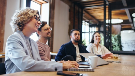 Vier Personen sitzen nebeneinander an einem langen Holztisch. Alle blicken in den rechten Bildbereich. Vor einer Person liegt ein Notizblock. Die Person neben ihr hat einen Laptop vor sich