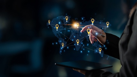 In der Mitte des Bildes ist eine Grafik der Kontinente der Welt. An manchen Stellen sind Icons für Lokalisierungen, die miteinander verbunden sind. Die rechte Hand einer Person zeigt mit einem Stift auf eine Stelle