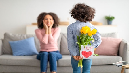 Kind steht mit dem Rücken zur Kamera und hält gelbe Tulpen sowie eine selbstgemachte Karte mit der Aufschrift „Mom“ hinter dem Rücken; im Hintergrund sitzt eine Person auf einem Sofa.