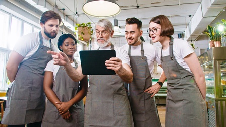 Frontale Aufnahme von fünf Personen mit Schürzen, die nebeneinander aufgereiht in einem Lokal stehen. Die mittlere Person hält ein Tablet, auf das alle blicken.