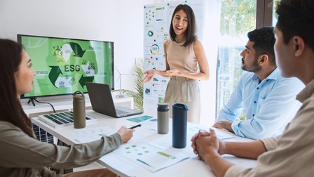 Drei Personen sitzen an einem Tisch. Eine Person steht neben dem Tisch. Auf dem Tisch sind ein Laptop und Zettel. Daneben ist ein großer Bildschirm, auf dem in der Mitte ESG steht. Daneben stehen Begriffe wie Environment, Social