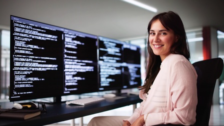 Porträt einer Person mit offenen, braunen Haaren, die an einem Schreibtisch sitzt und lächelt. Vor ihr auf dem Schreibtisch sind vier Bildschirme. Auf jedem Bildschirm ist ein HTMl-Code