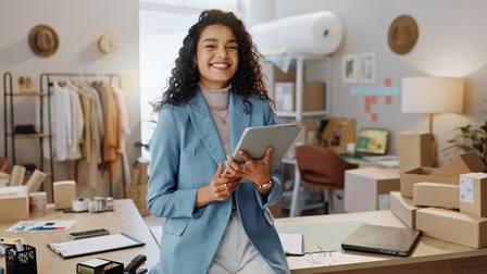 Lächelnde Person mit Tablet in Händen lehnt an Tisch, im Hintergrund verschwommen Arbeitsutensilien, gestapelte Kartonagen, Gestell mit hängenden Gewändern, Regale und Arbeitsplatz mit Computer