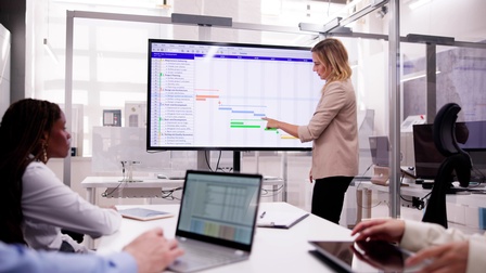 Eine Person steht seitlich neben einem großen Bildschirm. Auf dem Bildschirm sind verschieden farbige Balken, auf die sie deutet. Der Person gegenüber ist ein Tisch, an dem mehrere Personen sitzen, die zum Bildschirm blicken