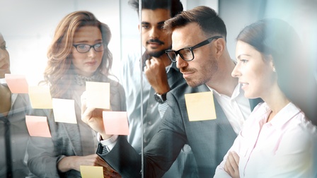 Gruppe von unterschiedlichen Personen steht vor einer Glaswand mit Post-Its und betrachtet diese