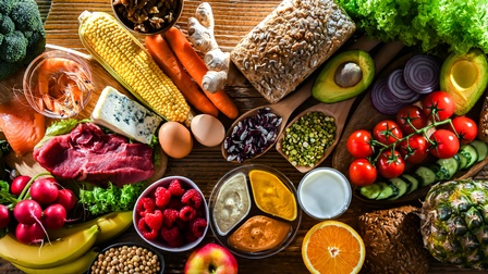 Topshot verschiedener Lebensmittel auf einer hölzernen Unterfläche, zum Beispiel Brot, Eier, Käse, Fleisch, Lachs, Bananen, Tomaten, Aufstriche, Samen, Avocados, Radieschen, Joghurt, Karotten, Ingwerwurzel