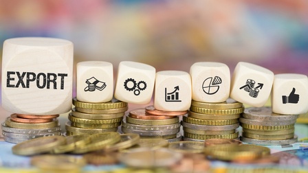 Euro-Münzen und Euro-Scheinen liegen auf einer Oberfläche, teilweise übereinander gestapelt. Auf den Münzstapeln liegen sieben Holzwürfel mit Symbolen zum Thema Finanzen, Wirtschaft, Export