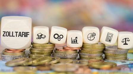 Nahaufnahme von fünf Münzstapeln nebeneinander. Auf den Münzen liegen sieben weiße Würfel. Auf dem ersten steht Zolltarif. Auf den anderen sind Symbole wie ein Taschenrechner oder eine Waage