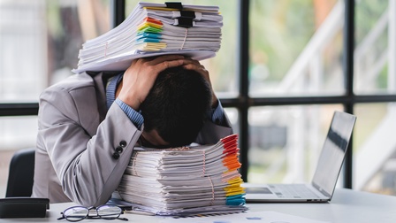 Eine Person in einem hellgrauen Sakko sitzt an einem Tisch und hat ihren Kopf auf einem hohen Stapel Papier abgelegt. Auf ihrem Kopf liegt ein weiterer Stapel Papiere. Neben ihr steht ein aufgeklappter Laptop