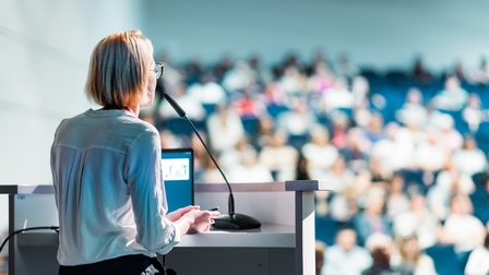 Person steht vor einem Pult mit Mikrofon, im Hintergrund sitzen Personen im Publikum